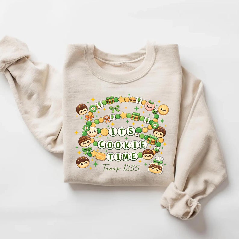 May include: Beige sweatshirt with the text "ITS COOKIE TIME" in a circle of cookies, stars, and gift boxes. The words "Troop 1235" are below the text. The sweatshirt has long sleeves and a crew neck.