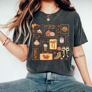 May include: Dark gray t-shirt featuring a collection of autumn-themed illustrations. The design includes pumpkins, pie, coffee, leaves, and the word "cozy". The shirt has a vintage, worn look.