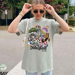 May include: A sage green short-sleeve t-shirt featuring a colorful graphic print of Disney characters including Mickey Mouse, Minnie Mouse, and Donald Duck in front of a castle, with the text Disneyland Est. 1955 printed below the characters.
