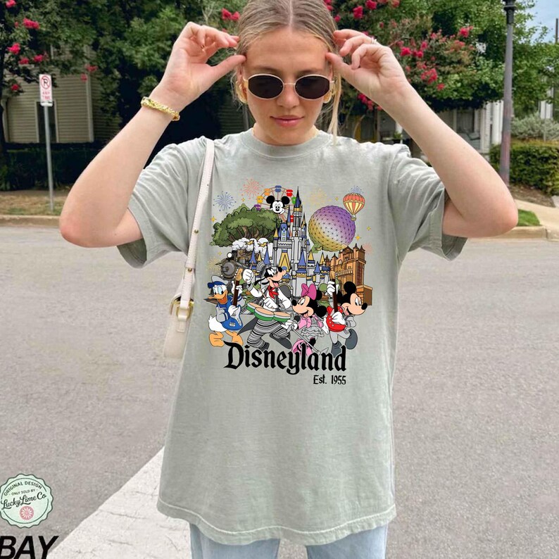 May include: A sage green short-sleeve t-shirt featuring a colorful graphic print of Disney characters including Mickey Mouse, Minnie Mouse, and Donald Duck in front of a castle, with the text Disneyland Est. 1955 printed below the characters.