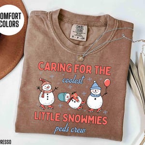 May include: Espresso-colored t-shirt with the text "CARING FOR THE coolest LITTLE SNOWMIES peds crew." The shirt features three cartoon snowmen wearing hats, scarves, and ice skates. A silver necklace is draped across the shirt.