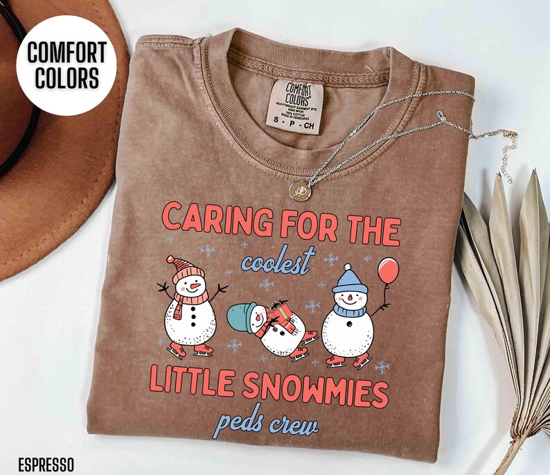 May include: Espresso-colored t-shirt with the text "CARING FOR THE coolest LITTLE SNOWMIES peds crew." The shirt features three cartoon snowmen wearing hats, scarves, and ice skates. A silver necklace is draped across the shirt.