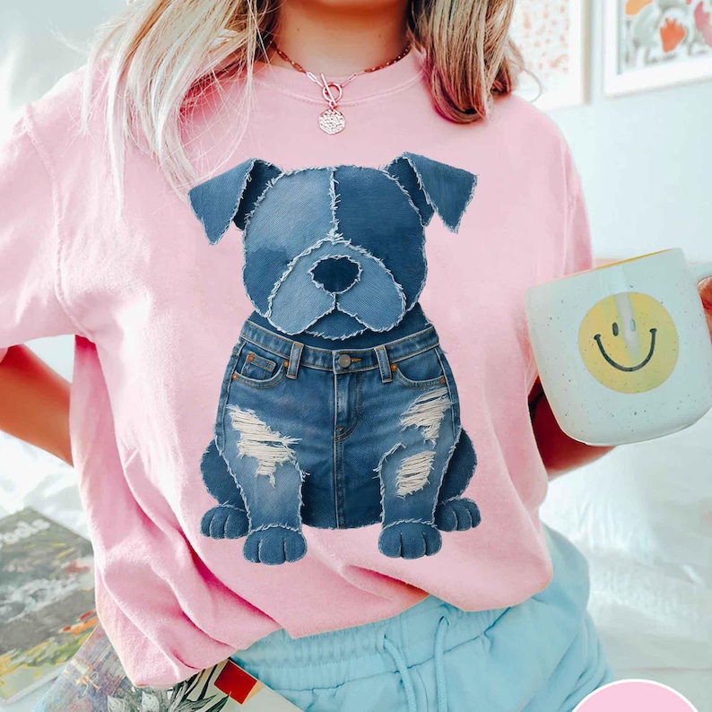 May include: A light pink t-shirt featuring a denim dog graphic. The dog is wearing denim jeans with distressed details. A white mug with a yellow smiley face is held in the background.