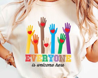 May include: A white t-shirt with a rainbow-colored design of hands reaching up with hearts in their palms. The text "EVERYONE is welcome here" is printed below the hands.