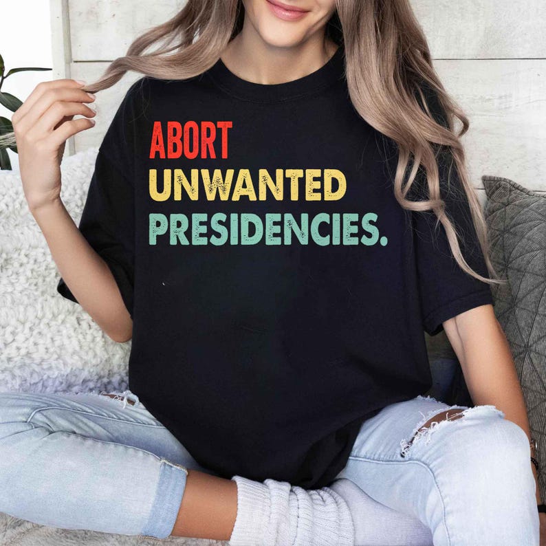 May include: Black t-shirt with the text "ABORT UNWANTED PRESIDENCIES" in red, yellow, and green. The shirt is a crew neck and is being worn.
