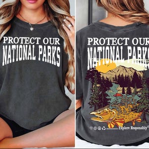 May include: A dark gray t-shirt with the text "Protect Our National Parks" on the front and a graphic of a mountain scene with a fish on the back. The back also includes the text "Explore Responsibly".