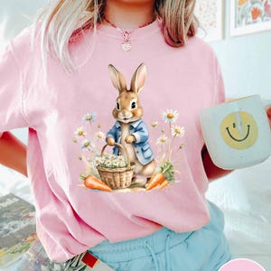 May include: A pink t-shirt with a cartoon bunny rabbit holding a basket of flowers. The bunny is wearing a blue jacket and is surrounded by daisies.