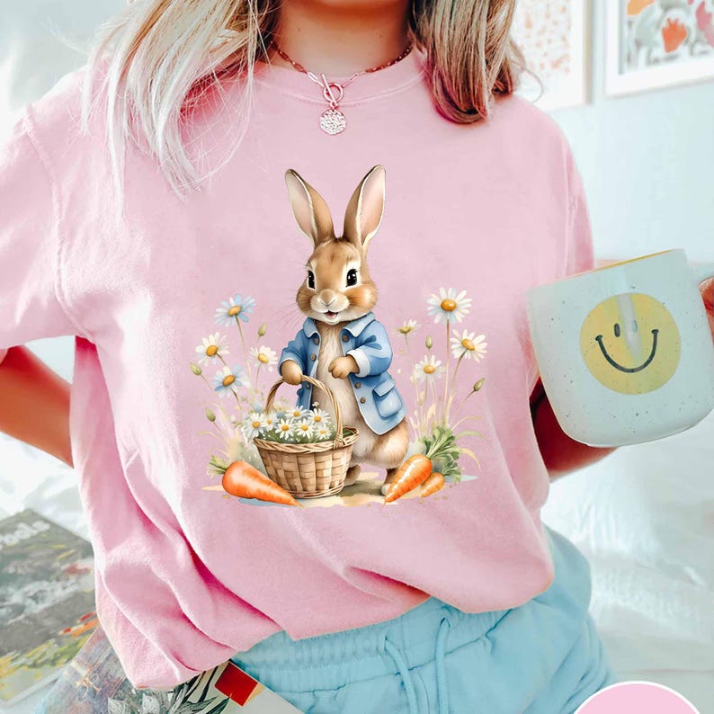 May include: A pink t-shirt with a cartoon bunny rabbit holding a basket of flowers. The bunny is wearing a blue jacket and is surrounded by daisies.