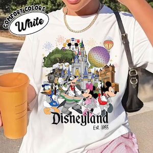 May include: A white t-shirt featuring a colorful graphic of Disney characters playing instruments in front of a castle, a geosphere, and a hot air balloon. The text reads Disneyland Est. 1955. The shirt includes a Comfort Colors White label graphic.