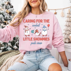 May include: A light pink sweatshirt with the text "CARING FOR THE coolest LITTLE SNOWMIES peds crew" and illustrations of three snowmen. One snowman wears a hat and scarf, another is lying down, and the third holds a balloon.