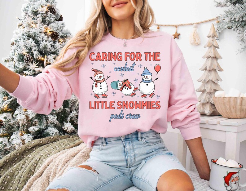 May include: A light pink sweatshirt with the text "CARING FOR THE coolest LITTLE SNOWMIES peds crew" and illustrations of three snowmen. One snowman wears a hat and scarf, another is lying down, and the third holds a balloon.