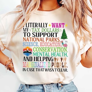 May include: White t-shirt with a colorful graphic print that reads "LITERALLY WANT MY $ TAX DOLLARS $ TO SUPPORT NATIONAL PARKS, SCIENCE, EDUCATION, CONSERVATION, MENTAL HEALTH, AND HELPING MARGINALIZED PEOPLE. IN CASE THAT WASN'T CLEAR."
