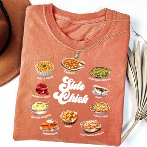 May include: A coral-colored t-shirt featuring illustrations of various side dishes, including mac and cheese, stuffing, and sweet potato casserole. The words "Side Chick" are printed in a retro font. A gold necklace is draped across the shirt.