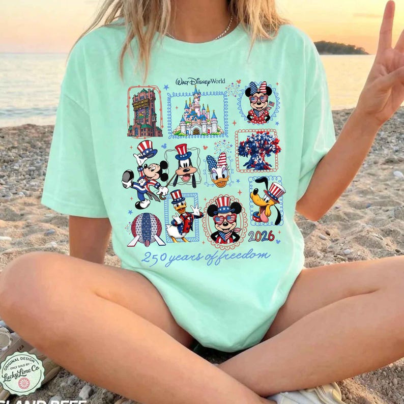 May include: A light mint green t-shirt featuring a graphic print of Walt Disney World icons and characters including Mickey Mouse, Minnie Mouse, Donald Duck, Goofy, and Pluto in patriotic red, white, and blue attire with the text 250 years of freedom.