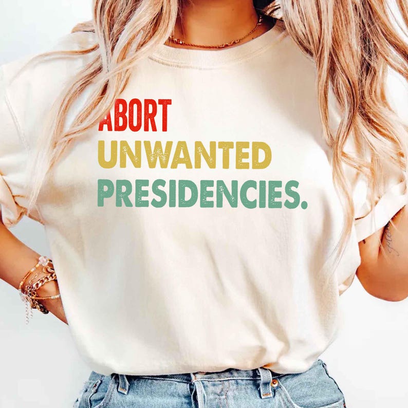 May include: Cream-colored t-shirt with the text "ABORT UNWANTED PRESIDENCIES" in red, yellow, and green block letters. The shirt has short sleeves and a relaxed fit.