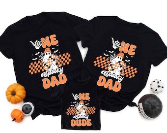 May include: Black t-shirts with a Halloween-themed design. The shirts feature the text "ONE Spooky DAD" and "ONE Spooky DUDE" with a ghost graphic, orange accents, and a checkered pattern. Decorative pumpkins and Halloween-themed balls are also present.