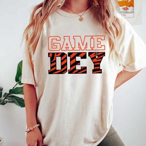 May include: A white t-shirt with the text "GAME DAY" in black and orange letters. The "DAY" is in a tiger stripe pattern.