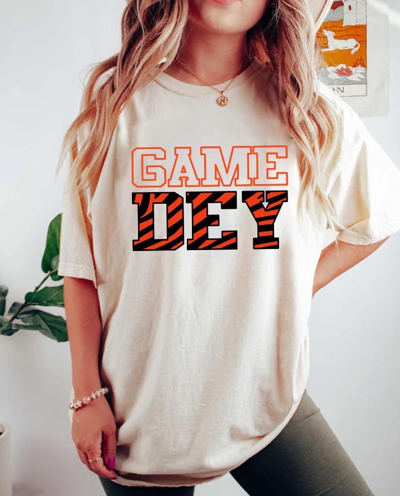 May include: A white t-shirt with the text "GAME DAY" in black and orange letters. The "DAY" is in a tiger stripe pattern.