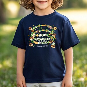 May include: Navy blue t-shirt with a colorful graphic that says "It's Cookie Time" with cookie and star illustrations. The text "Troop 1235" is also included in the design. The shirt is worn by a child.