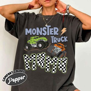Puede incluir: Camiseta gris oscuro con un dise&ntilde;o gr&aacute;fico de dos monster trucks y el texto "MONSTER TRUCK MAMA". La palabra "MAMA" tiene un patr&oacute;n de cuadros. La camiseta tambi&eacute;n tiene las palabras "COMFORT COLORS" y "Pepper" impresas.