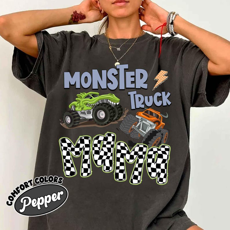 Puede incluir: Camiseta gris oscuro con un dise&ntilde;o gr&aacute;fico de dos monster trucks y el texto "MONSTER TRUCK MAMA". La palabra "MAMA" tiene un patr&oacute;n de cuadros. La camiseta tambi&eacute;n tiene las palabras "COMFORT COLORS" y "Pepper" impresas.