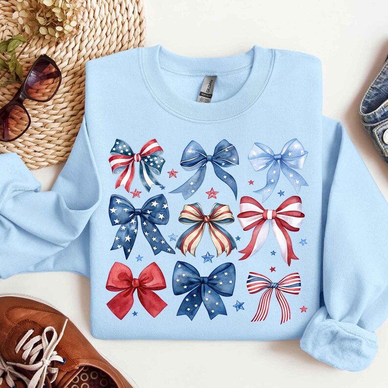 May include: A light blue sweatshirt with a design of red, white, and blue bows. The bows are arranged in a grid pattern and have a watercolor effect. The sweatshirt is a comfortable and stylish option for everyday wear.