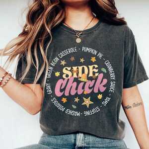 May include: Dark gray t-shirt with a circular design featuring the text "SIDE Chick" in pink and yellow, surrounded by Thanksgiving food names and autumn leaves. The shirt has a vintage, worn look.