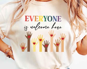 May include: A white t-shirt with the text "EVERYONE is welcome here" in rainbow colors. The text is surrounded by a row of raised hands with hearts in the palms, representing different skin tones.