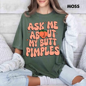 Op de afbeelding: Mosgroen t-shirt met de tekst "ASK ME ABOUT MY BUTT PIMPLES" in oranje letters. Een perzikafbeelding staat in het midden. Het woord "MOSS" is rechtsboven te zien.
