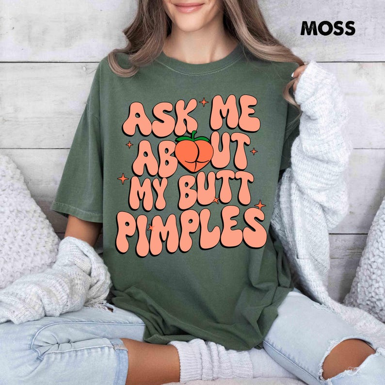 Op de afbeelding: Mosgroen t-shirt met de tekst "ASK ME ABOUT MY BUTT PIMPLES" in oranje letters. Een perzikafbeelding staat in het midden. Het woord "MOSS" is rechtsboven te zien.