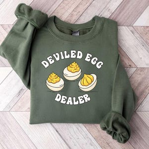May include: An olive green sweatshirt with the words "DEVILED EGG DEALER" in white, along with an illustration of three deviled eggs. The eggs have a yellow filling and are on a wooden surface.