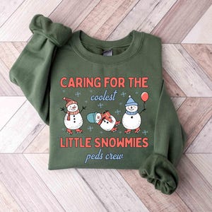 May include: An olive green sweatshirt with the text "CARING FOR THE coolest LITTLE SNOWMIES peds crew". The graphic features three snowmen wearing hats and scarves, ice skating, and holding a balloon. The sweatshirt is folded on a wooden surface.