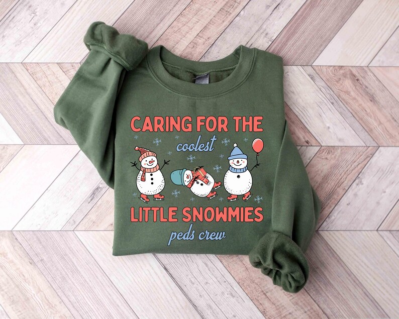 May include: An olive green sweatshirt with the text "CARING FOR THE coolest LITTLE SNOWMIES peds crew". The graphic features three snowmen wearing hats and scarves, ice skating, and holding a balloon. The sweatshirt is folded on a wooden surface.