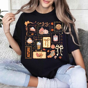 May include: Black t-shirt with a colorful autumn-themed graphic. The design includes pumpkins, a slice of pie, a sweater, and the word "cozy" in script. The word "Autumn" is also featured.