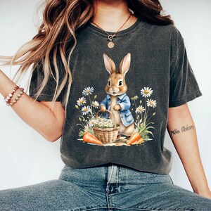 May include: A dark gray t-shirt with a vintage look featuring a cartoon bunny holding a basket of flowers and carrots. The bunny is wearing a blue jacket.