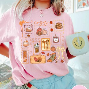 May include: Pink t-shirt with a colorful autumn-themed graphic. The design includes pumpkins, leaves, a sweater, and the word "cozy." A white mug with a yellow smiley face is held in the hand.