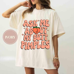 Op de afbeelding: Ivoor kleurig t-shirt met de tekst "ASK ME ABOUT MY BUTT PIMPLES" in een retro lettertype. Een perzikafbeelding staat boven de tekst. Het shirt heeft korte mouwen en een relaxte pasvorm.