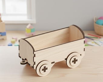 Op de afbeelding: Een houten speelgoedwagen met een lichtbruine afwerking. De wagen heeft een rechthoekige behuizing met afgeronde randen en twee wielen aan elke kant, elk met een kruisontwerp. De wagen is ontworpen voor speelgoedopslag.