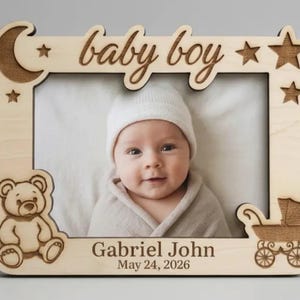 Puede incluir: Marco de fotos de madera para bebé con las palabras "baby boy" grabadas. El marco presenta una luna, estrellas, un oso de peluche y un cochecito. También están grabados el nombre y la fecha de nacimiento del bebé.