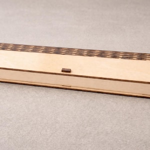Könnte beinhalten: Eine rechteckige Holzkiste mit hellem Braunton. Der Deckel hat ein dekoratives, dunkelbraunes, geometrisches Muster. Die Kiste hat einen kleinen, rechteckigen Verschluss und ist zur Aufbewahrung gedacht. Die Kiste ist ca. 25 cm lang.