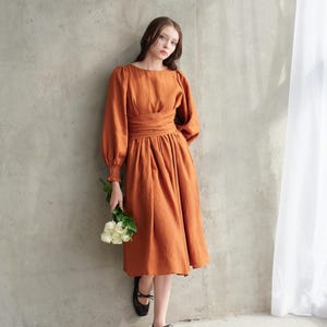 Puede incluir: Un vestido de color naranja-marrón con mangas largas y cintura ceñida. El vestido tiene un escote barco y una falda fluida. La modelo lleva bailarinas negras y sostiene un ramo de flores blancas. El vestido está hecho de una tela suave y fluida.