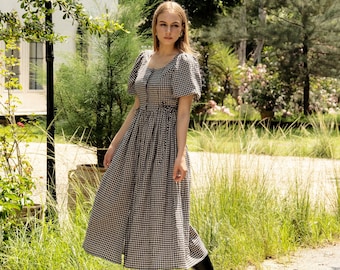 May include: A woman wearing a black and white gingham midi dress with puffed sleeves and black knee-high boots. She is standing on a path in a garden setting.
