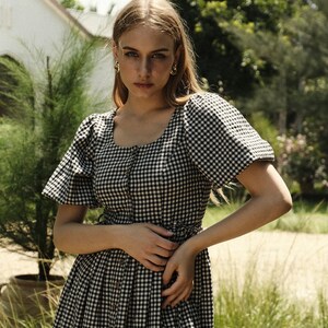 Puede incluir: Una mujer con un vestido de cuadros negros y blancos con mangas abullonadas. El vestido tiene un escote cuadrado y botones en la parte delantera. Est&aacute; de pie en un entorno de jard&iacute;n.