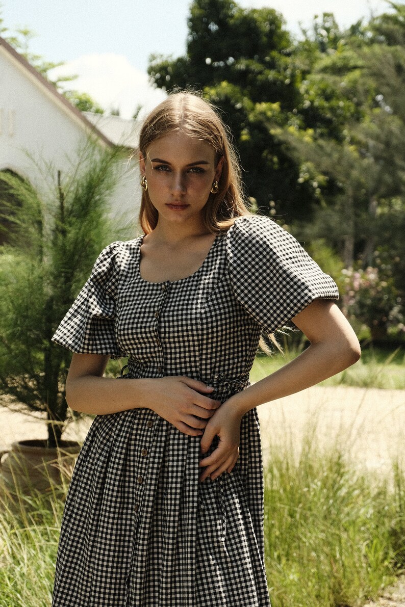 Puede incluir: Una mujer con un vestido de cuadros negros y blancos con mangas abullonadas. El vestido tiene un escote cuadrado y botones en la parte delantera. Est&aacute; de pie en un entorno de jard&iacute;n.