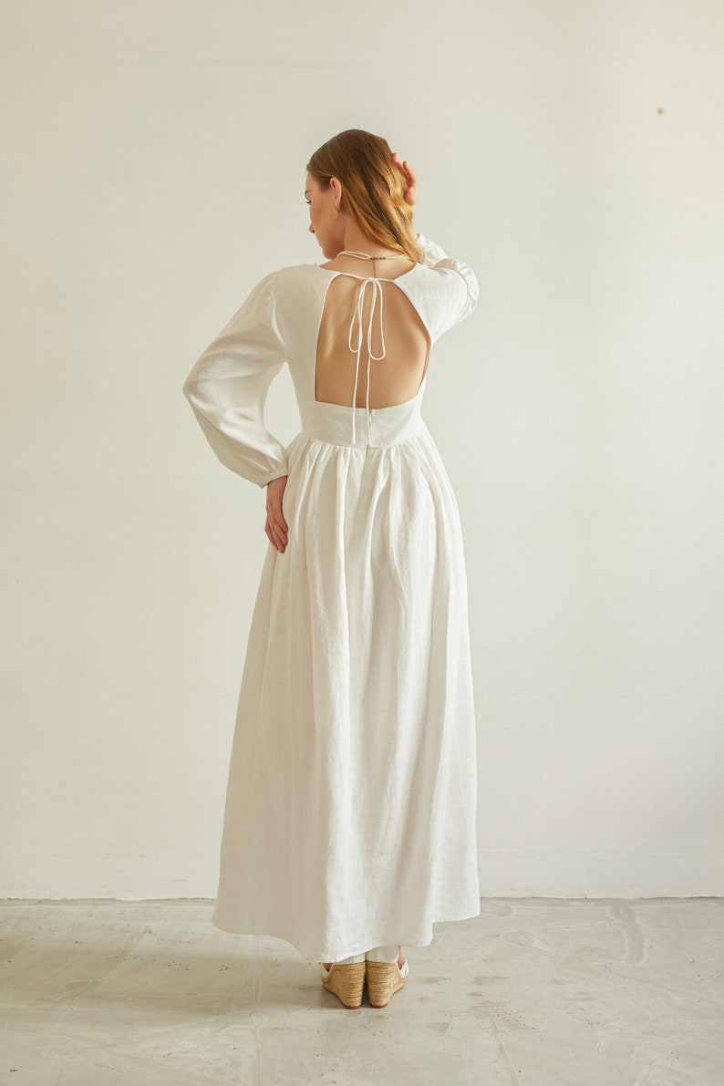 May include: A white maxi dress with long sleeves and a low back with a tie detail. The dress is made of a lightweight fabric and has a flowing silhouette.