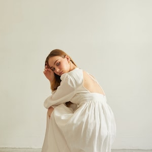 May include: A woman wearing a white linen dress with a low back and long sleeves. She is sitting on the floor with her head resting on her hand.