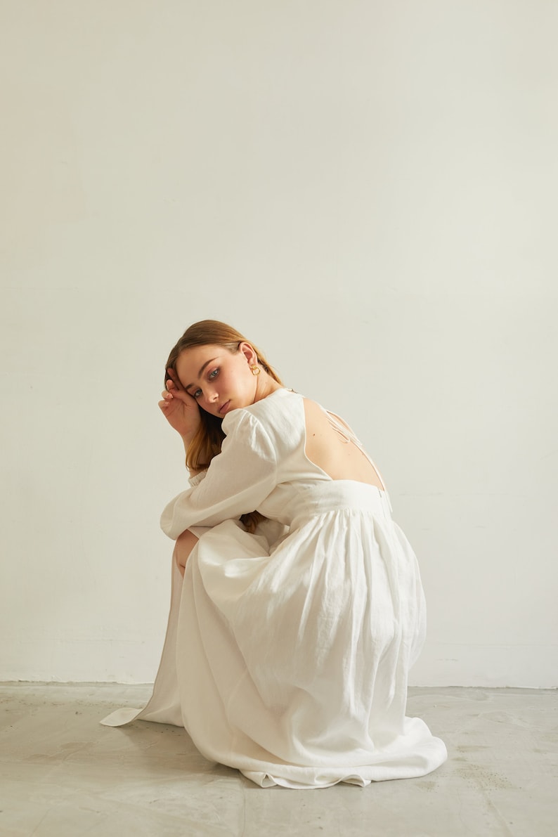 May include: A woman wearing a white linen dress with a low back and long sleeves. She is sitting on the floor with her head resting on her hand.