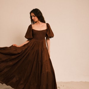 Pode incluir: Uma mulher vestindo um vestido maxi marrom em camadas com mangas bufantes. O vestido tem um decote quadrado e um corpete ajustado. Ela est&aacute; em p&eacute; em uma superf&iacute;cie arenosa.