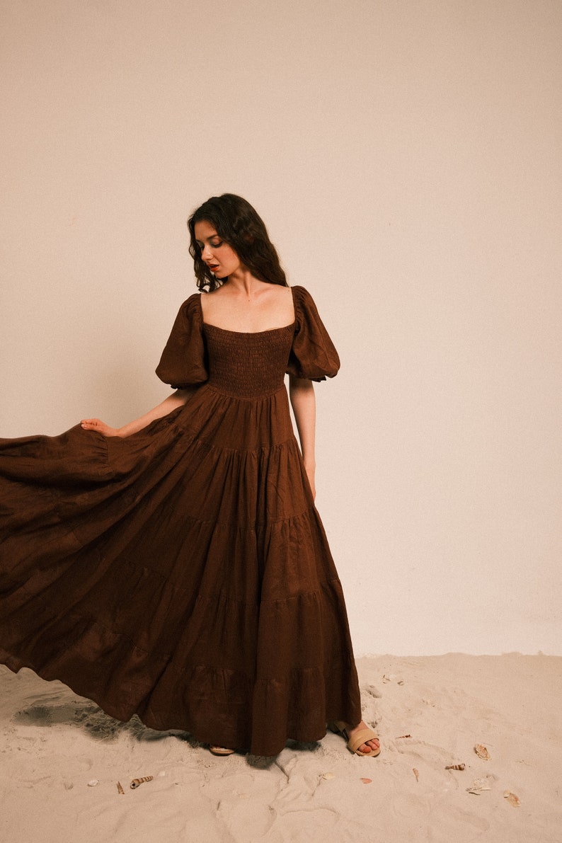Pode incluir: Uma mulher vestindo um vestido maxi marrom em camadas com mangas bufantes. O vestido tem um decote quadrado e um corpete ajustado. Ela est&aacute; em p&eacute; em uma superf&iacute;cie arenosa.