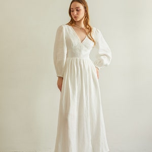 May include: A white linen maxi dress with a V-neckline, long sleeves, and a gathered skirt. The dress has a delicate lace trim around the neckline.